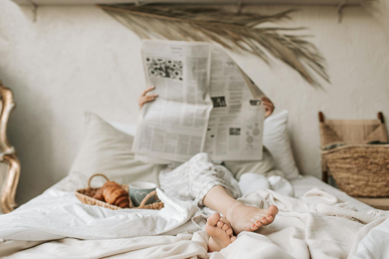 Person relaxing in bed with a newspaper, coffee, and croissant for a calm morning vibe.